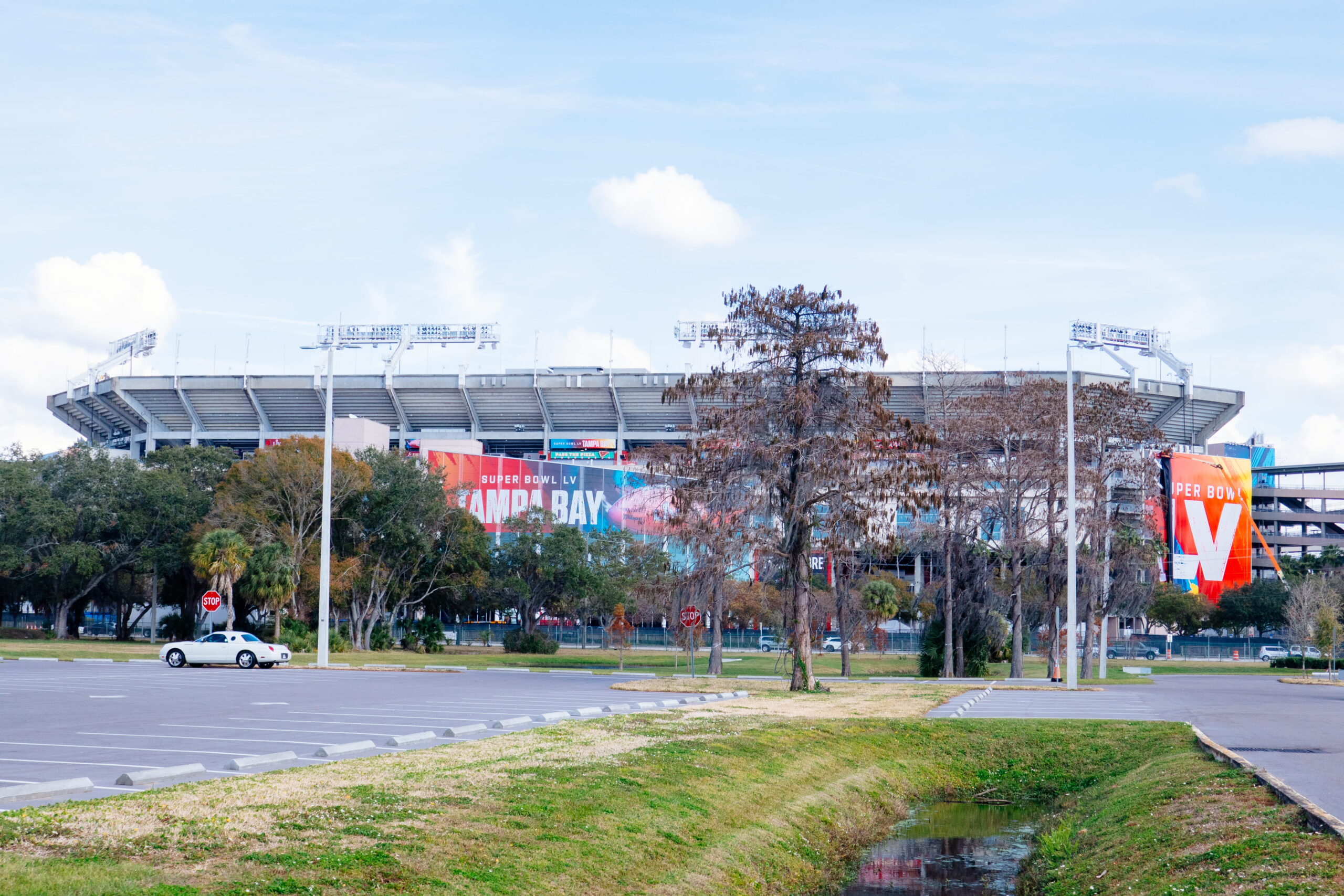 Raymond James Stadium exterior in Tampa, Florida, viewed from a nearby parking lot with trees, light poles, and Super Bowl LV signage visible on the stadium facade.